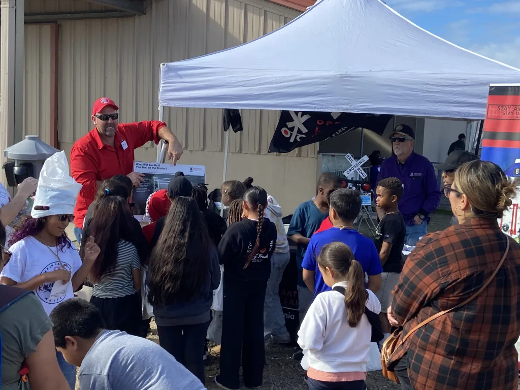 A group of children gathers around a booth under a white canopy tent, listening to two men, one in a red shirt and cap, explaining a display about railway safety. The event is outdoors near a building on a sunny day.