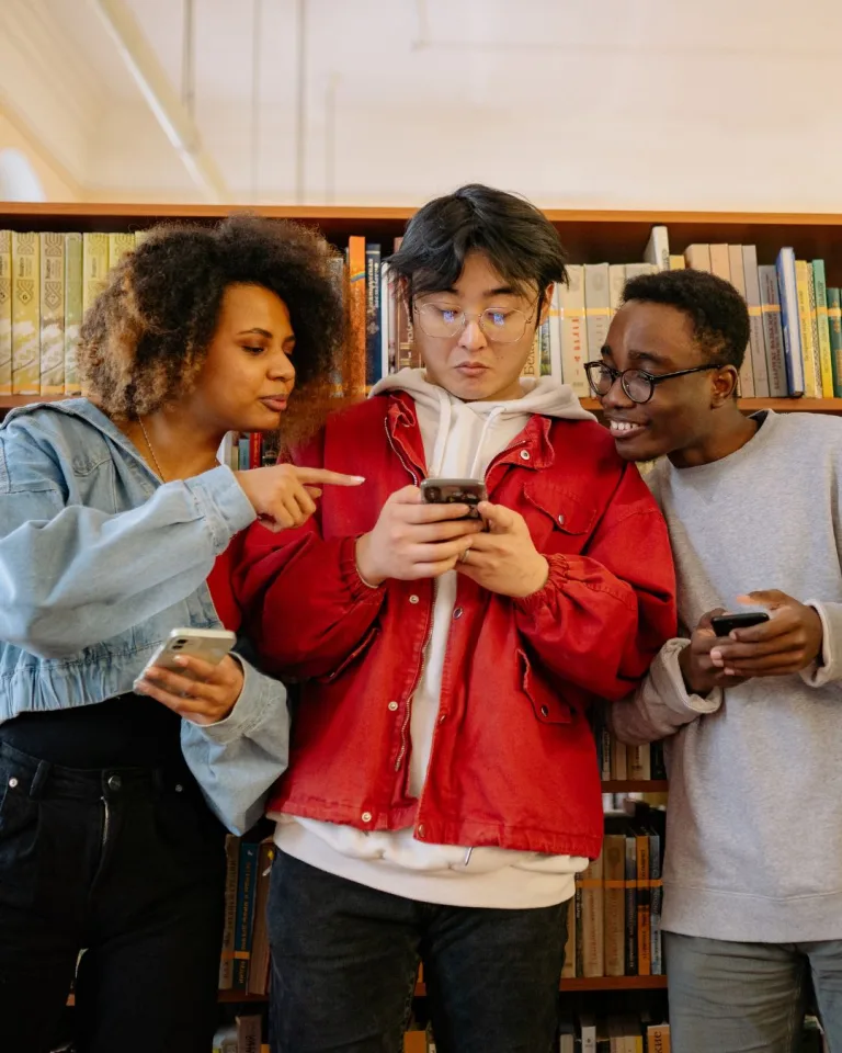 Three young adults stand in front of bookshelves. The person in the red jacket checks their phone as the others smile and point—each sharing rail safety awareness on social media from their own devices.