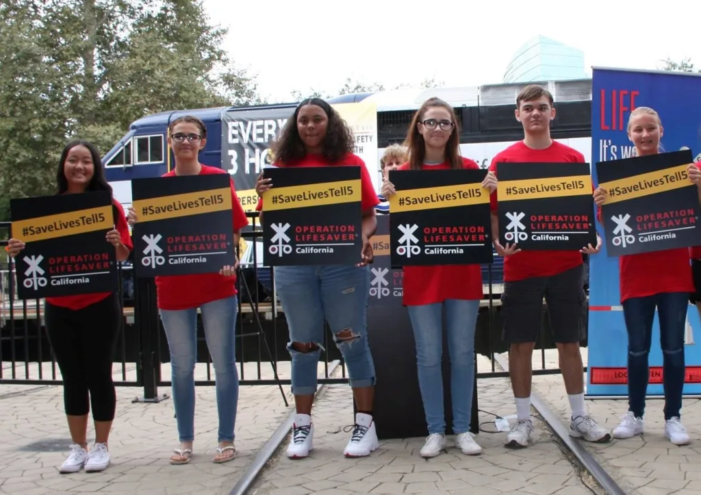 Six young people stand on train tracks, each holding #SaveLivesTell5 signs promoting Operation Lifesaver California, with a train, banners, and trees visible in the background.